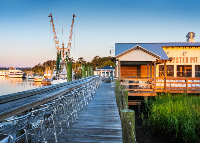 A long wooden pier with rows of metal chairs leads to The Oyster Pit restaurant on the water at sunset, with shrimp boats docked nearby.