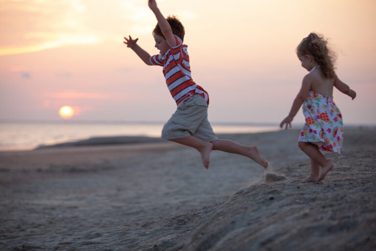 Young brother and sister play as they run and jump down a sand dune on the beach at sunset.