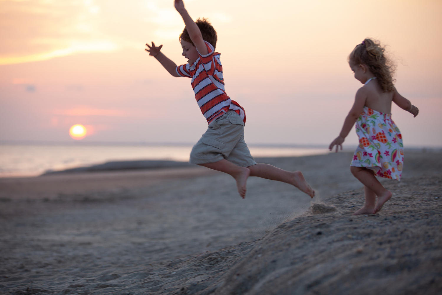 Young brother and sister play as they run and jump down a sand dune on the beach at sunset.